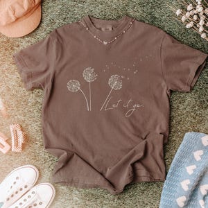 May include: A brown t-shirt with a dandelion graphic and the words "Let it go" in a cursive font. The shirt is surrounded by accessories, including a peach-colored hat, white sneakers, and hair clips.
