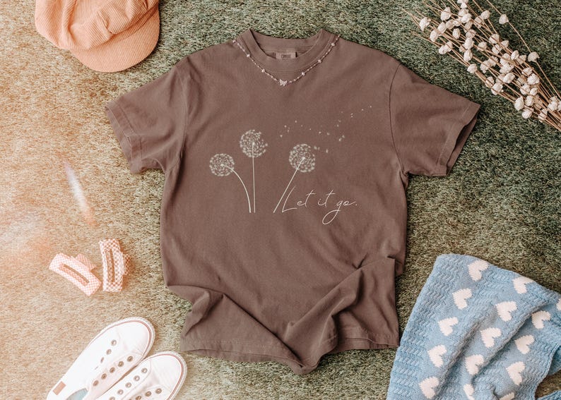 May include: A brown t-shirt with a dandelion graphic and the words "Let it go" in a cursive font. The shirt is surrounded by accessories, including a peach-colored hat, white sneakers, and hair clips.