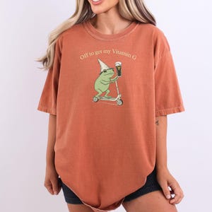 May include: Burnt orange t-shirt with the phrase "Off to get my Vitamin G." The graphic features a green frog wearing a hat and riding a scooter while holding a beer. The shirt has short sleeves and a relaxed fit.