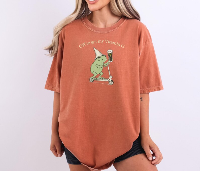 May include: Burnt orange t-shirt with the phrase "Off to get my Vitamin G." The graphic features a green frog wearing a hat and riding a scooter while holding a beer. The shirt has short sleeves and a relaxed fit.