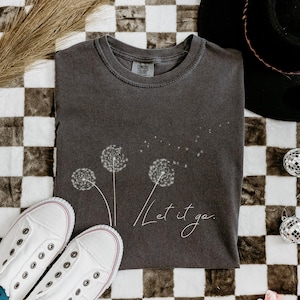 May include: Dark gray t-shirt with a dandelion design and the words "Let it go." White canvas sneakers and decorative disco balls are also in the image. The shirt is folded on a checkered surface.