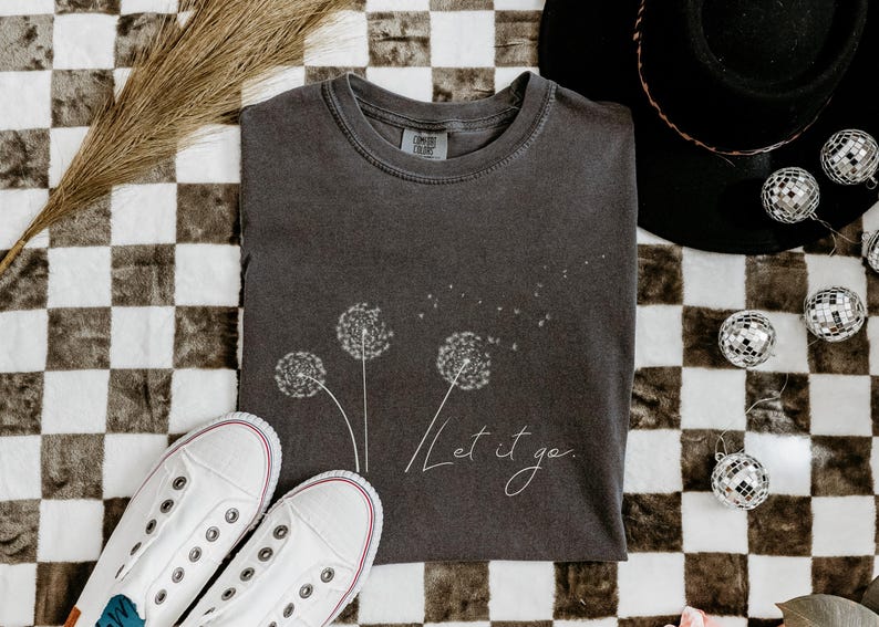 May include: Dark gray t-shirt with a dandelion design and the words "Let it go." White canvas sneakers and decorative disco balls are also in the image. The shirt is folded on a checkered surface.