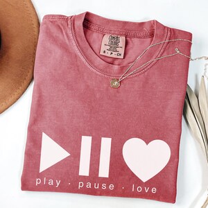May include: A dusty rose-colored t-shirt with a white graphic of a play button, pause bars, and a heart. The words "play, pause, love" are printed below. A silver necklace with a pendant and a brown hat are also visible.