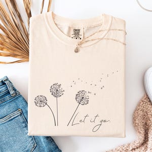 May include: Cream-colored t-shirt with a dandelion graphic and the words "Let it go." The shirt is paired with blue jeans, white sneakers, and a necklace. A dried palm leaf adds a natural touch to the flat lay.