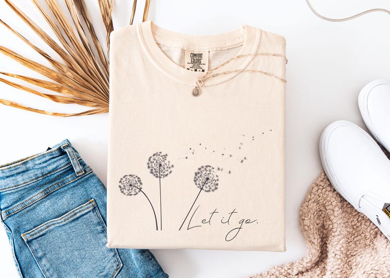 May include: Cream-colored t-shirt with a dandelion graphic and the words "Let it go." The shirt is paired with blue jeans, white sneakers, and a necklace. A dried palm leaf adds a natural touch to the flat lay.