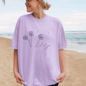 May include: A light purple t-shirt featuring a dandelion design with the words "Let it go" in a handwritten font. The design includes three dandelion flowers with seeds blowing away. The shirt is a soft, comfortable material.