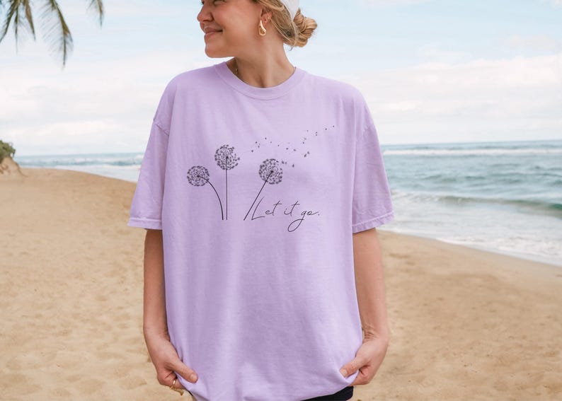 May include: A light purple t-shirt featuring a dandelion design with the words "Let it go" in a handwritten font. The design includes three dandelion flowers with seeds blowing away. The shirt is a soft, comfortable material.