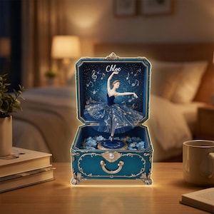 May include: A teal music box with a ballerina figure in a blue dress with star detailing. The box has a handle and decorative features. The name "Chloe" is written on the top. The interior is decorated with blue flowers and musical notes.