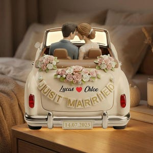 Puede incluir: Un adorno para tarta de boda de color crema con forma de coche descapotable vintage. El coche presenta una figura de novios, arreglos florales y una pancarta que dice "Just Married". Se muestra la fecha "14.07.2025".