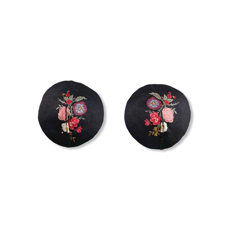 Flat lay of lingerie silk floral pasties in black satin with multi-colored floral bouquet print in circle shape with straight black stitch line. Satin has a muted shine and a romantic Victorian feel with purple, pink, red, green, and yellow hues.