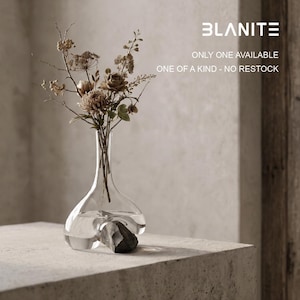 May include: A clear glass decanter with a wide, rounded base and a narrow neck, containing a dark grey stone inside the base. The decanter holds a bouquet of dried brown flowers. Text on the image reads BLANITE, ONLY ONE AVAILABLE, ONE OF A KIND - NO RESTOCK.