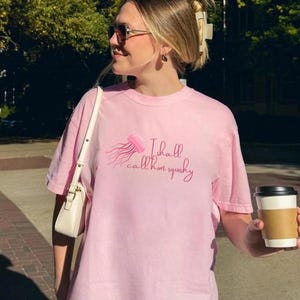 May include: A light pink t-shirt with a jellyfish graphic and the text "I shall call him squishy". The person is holding a white purse and a coffee cup. The background includes trees and a building.