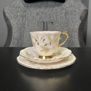 Puede incluir: Un juego de té vintage con una taza, un platillo y un plato de postre. Las piezas de porcelana blanca presentan ribetes dorados y un delicado diseño de hojas doradas y verdes. El interior de la taza es rosa.