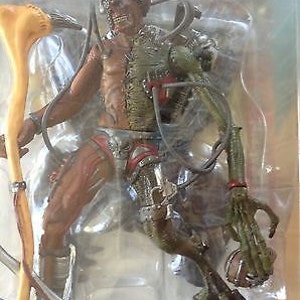 May include: Action figure of a cyborg-like character with exposed flesh and mechanical components. The figure has a skeletal face, brown and green coloring, and is holding a scythe-like weapon. The figure is packaged in a clear plastic container.
