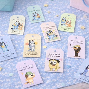 以下が含まれることがあります： 漫画の犬のイラストとテキストが描かれた、カラフルなギフトタグのコレクション。タグには「ありがとう」や「来てくれて嬉しい」などのフレーズが書かれています。青い模様の表面に散らばっており、パーティーやお祝いのテーマを示唆しています。