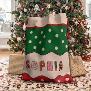 May include: A large Christmas gift sack with a green polka dot pattern and the name "SOPHIA" in colorful letters. The sack has a red drawstring and is decorated with red and white trim. A Christmas tree is in the background.
