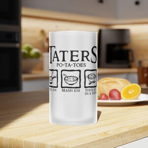 May include: A white frosted glass with the words "TATERS PO•TA•TOES" in black, along with three illustrated steps: boil em, mash em, and stick em in a stew. The glass is on a wooden surface.