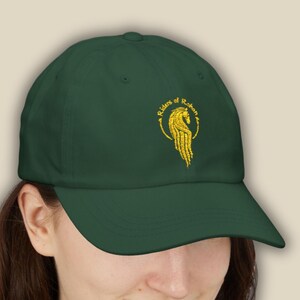 May include: A dark green baseball cap featuring the text "Riders of Rohan" in a gold, stylized font. The cap has a curved brim and a gold emblem of a horse. The hat is made of a soft material.