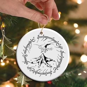 May include: White ceramic Christmas ornament featuring a black illustration of a mountain, a flying creature, and text in a circular pattern. The ornament is held by a gold string and hangs in front of a Christmas tree.