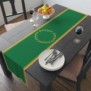 May include: A green table runner with a gold border and a gold ring design in the center. The runner is on a dark wood table set with plates, silverware, wine glasses, and a bowl of fruit. A decorative piece for a dining table.
