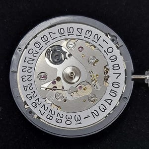 Pode incluir: Close-up de um movimento de relógio com uma estrutura de metal cinza e um mostrador branco exibindo os dias do mês. Os intrincados mecanismos internos são visíveis, juntamente com dois pequenos componentes de metal e uma ferramenta.