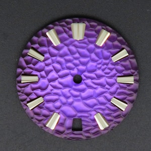 Pode incluir: Um mostrador de relógio roxo com um padrão texturizado e orgânico. Possui doze marcadores de hora retangulares prateados. O centro tem um pequeno orifício escuro. O design é circular.