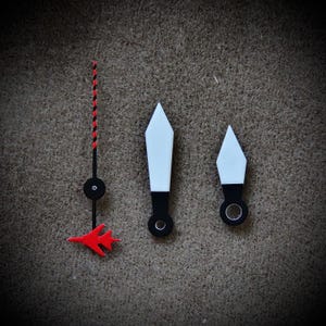 May include: A collection of watch hands, including a red and black striped second hand with a red airplane design, and two white and black hands. The hands have a black base and a white arrow-shaped tip.