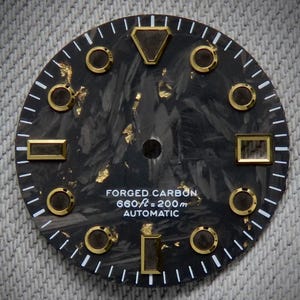 May include: Close-up of a watch face with a black forged carbon background and gold-colored accents. The face features a variety of shapes, including circles, rectangles, and a triangle. White text reads "FORGED CARBON 660 ft = 200 m AUTOMATIC."
