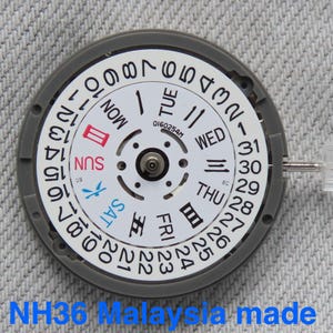 Puede incluir: Un componente circular de movimiento de reloj con una esfera blanca y números negros del 1 al 31 en el borde. El centro muestra los días de la semana en inglés y kanji, con el domingo en rojo y el sábado en azul. Texto NH36 Malaysia made.