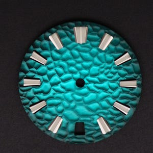 May include: A teal watch face with a textured surface and silver hour markers. The watch face is circular with a small central hole and a square opening at the bottom. The watch face has a diameter of approximately 3.8 cm.