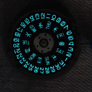 Puede incluir: Un disco de calendario de reloj circular con fondo negro y números cian brillantes del 1 al 31, además de los días de la semana en inglés y japonés. El centro presenta un componente de engranaje mecánico con varios orificios circulares pequeños.