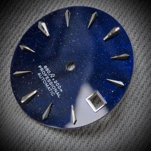 Puede incluir: Esfera de reloj circular con un fondo azul oscuro moteado, que recuerda a un cielo estrellado. Marcadores de hora triangulares plateados y un marcador cuadrado a las 3 en punto. El texto "660 ft = 200m PROFESSIONAL AUTOMATIC" está impreso en la esfera.