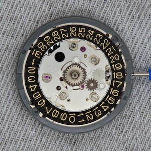 May include: Close-up of a watch movement with a black and gold date dial. The dial features numbers 1 through 31. The watch mechanism includes gears, screws, and jewels. A blue tool is inserted into the mechanism.