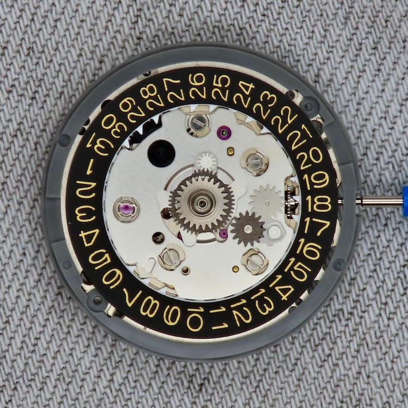 May include: Close-up of a watch movement with a black and gold date dial. The dial features numbers 1 through 31. The watch mechanism includes gears, screws, and jewels. A blue tool is inserted into the mechanism.