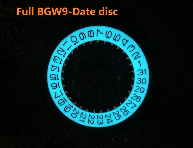 May include: A glowing blue watch date disc with white numerals against a black background. The disc is circular with numbers 1-31 around the edge. The text "Full BGW9-Date disc" is at the top.