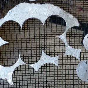 May include: A collection of silver watch faces and cutouts, some with etched patterns, arranged on a black mesh surface. The watch faces have various hole patterns and markings.