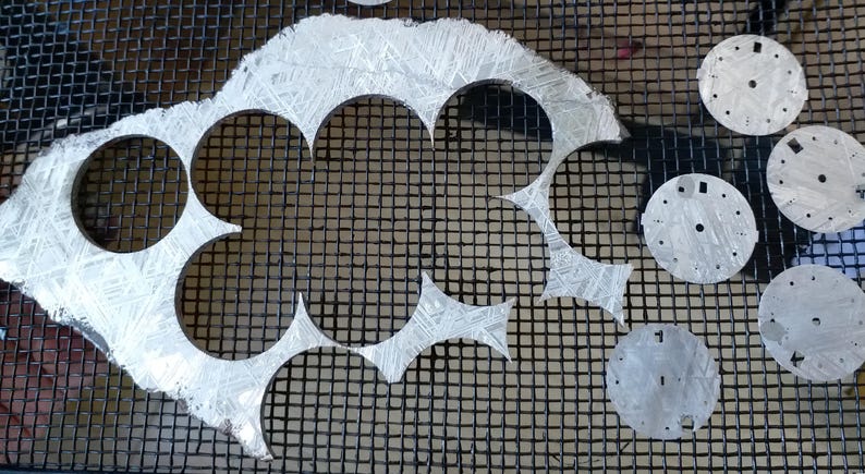 May include: A collection of silver watch faces and cutouts, some with etched patterns, arranged on a black mesh surface. The watch faces have various hole patterns and markings.