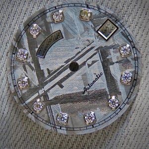 May include: A watch face with a textured gray background and a circular design. It features twelve diamond-like markers around the perimeter and a diamond-shaped marker. The word "Automatic" is written in cursive. The watch face has a diameter of approximately 1.5 inches.