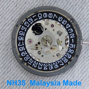 May include: A watch movement with a grey frame and a black dial displaying white numbers. The internal mechanisms are visible. The text "NH35 Malaysia Made" is at the bottom of the image.
