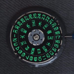 May include: A circular black watch movement calendar disc featuring luminous green numerals from 1 to 31 and days of the week in both English and Japanese characters arranged in concentric rings around a central metallic gear mechanism.