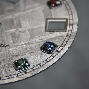 May include: Close-up of a watch face with a textured, gray surface and several colorful gemstones. The gemstones are square-cut and set in silver bezels. A rectangular silver frame is also visible.
