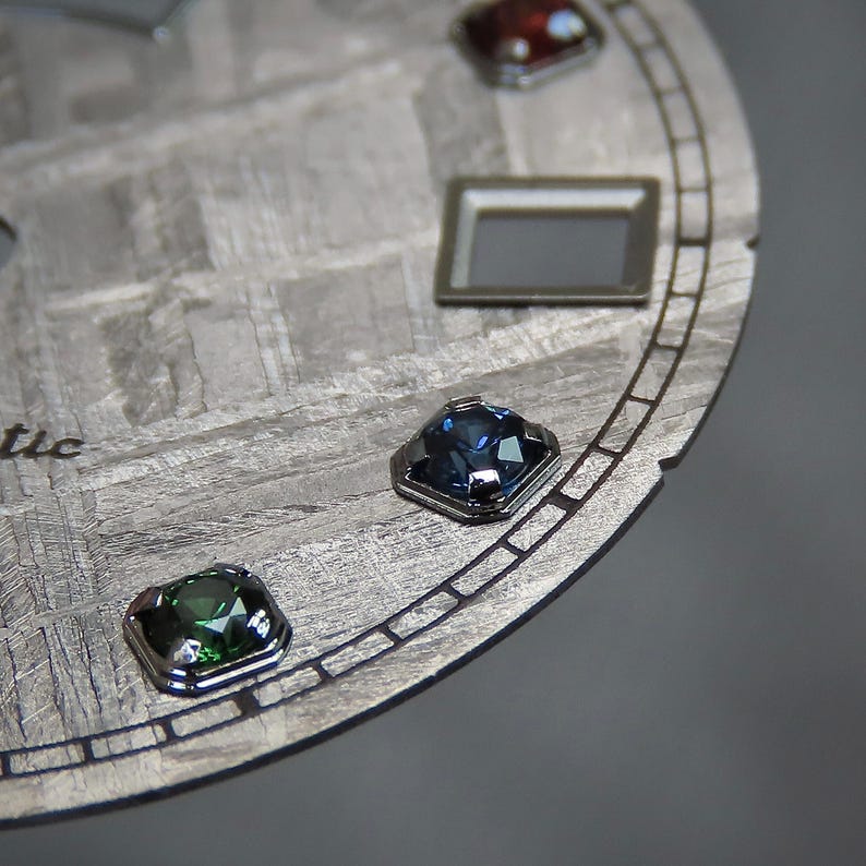 May include: Close-up of a watch face with a textured, gray surface and several colorful gemstones. The gemstones are square-cut and set in silver bezels. A rectangular silver frame is also visible.