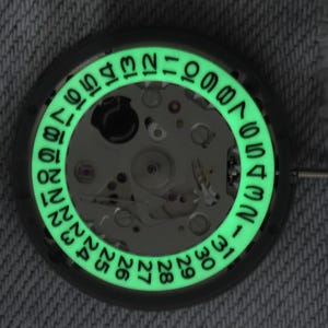 May include: A close-up of a watch movement featuring a luminous green date ring. The ring displays numbers from 1 to 31, indicating the days of the month. The watch mechanism is visible within the ring.