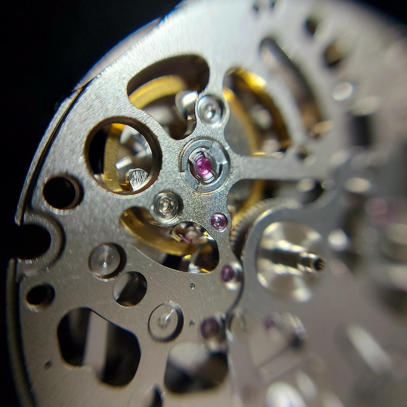 May include: Close-up of a watch mechanism with intricate details. The image showcases silver-colored metal components, including gears, screws, and jewels. The design features circular and linear elements, with a focus on precision and craftsmanship. The background is dark.
