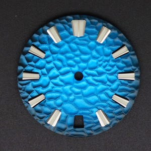 May include: A circular watch face with a textured, blue surface and silver hour markers. The face has a central hole and a rectangular opening at the bottom. The design is modern and stylish.