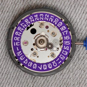May include: Close-up of a watch movement with a purple date ring. The watch mechanism features silver gears, screws, and jewels. The date ring displays numbers from 1 to 31 in white font. A blue tool is visible on the right side.