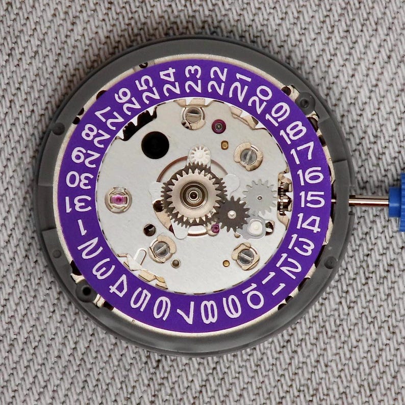 May include: Close-up of a watch movement with a purple date ring. The watch mechanism features silver gears, screws, and jewels. The date ring displays numbers from 1 to 31 in white font. A blue tool is visible on the right side.