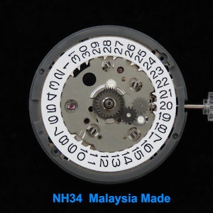 May include: Close-up of a watch movement with a white date ring displaying numbers. The movement has various gears and components, with the text "NH34 Malaysia Made" at the bottom. The watch movement is set against a black background.