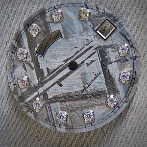 May include: A watch face with a textured gray background and a circular design. It features twelve diamond-like markers around the perimeter and a diamond-shaped marker. The word "Automatic" is written in cursive. The watch face has a diameter of approximately 1.5 inches.