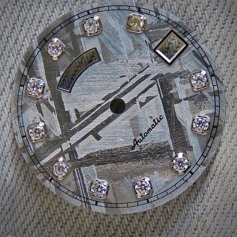 May include: A watch face with a textured gray background and a circular design. It features twelve diamond-like markers around the perimeter and a diamond-shaped marker. The word "Automatic" is written in cursive. The watch face has a diameter of approximately 1.5 inches.